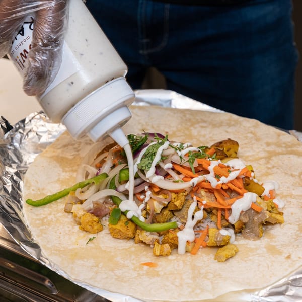 A hand in a glove squeezes white sauce onto a WOW TIKKA Indian wrap filled with chicken, rice, and vegetables.
