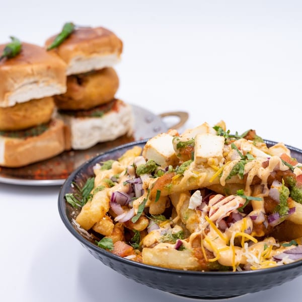 A black bowl of loaded Indian fries with paneer, sauces, and onions, with vada pav in the background.